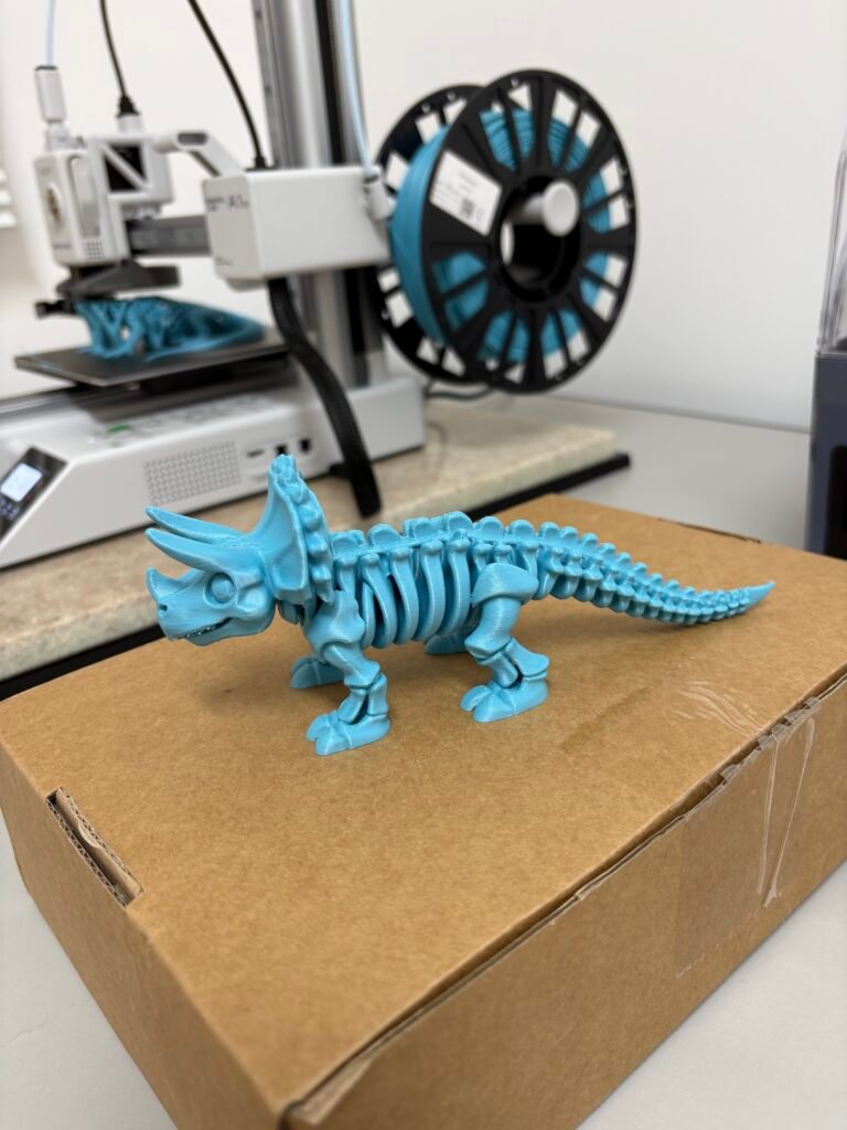 Triceratops impresso em 3D sendo finalizado na impressora, ao lado do rolo de filamento azul.